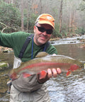 Angler holding a large trout in a forest stream, showcasing freshwater fishing success