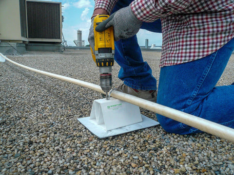 White 6-inch PVC rooftop pipe support block on a gravel roof with a pipe resting and a worker drilling.