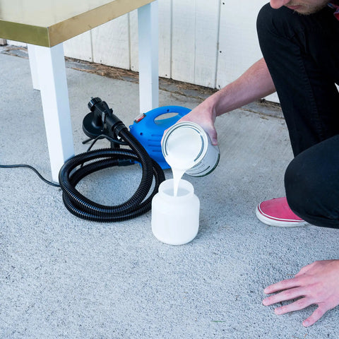 Person pouring paint into One Touch Paint Sprayer for home DIY projects, blue tool next to table on concrete surface.