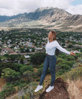 Woman standing on mountain, overlooking town, wearing white top and patterned leggings with mountains in background.