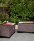 Two Ravenna 133 Gallon Water Resistant Deck Boxes For Outdoor Storage in a garden, filled with pillows and blankets, brown design.