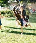 Children playing in backyard with water guns, one child being splashed while sitting on a zip line, grass and trees in background.