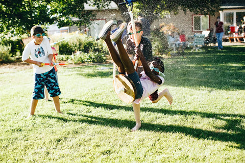 Children playing in backyard with water guns, one child being splashed while sitting on a zip line, grass and trees in background.