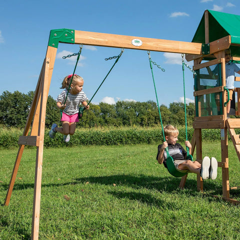 Two kids playing on the Buckley Hill Cedar Wood Swing Set With Wave Slide, Elevated Play Deck, Rock Wall Ladder, Belt Swings, Canopy Roof.