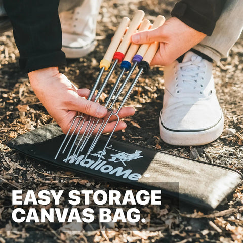 Person holding 5-pack of long 32 inch smores sticks kit with stainless steel skewers and smooth wooden handles for marshmallow roasting at the campfire fire pit gear for outdoor camping durable essentials with canvas storage bag on ground.