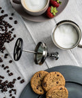 Handheld Milk Frother For Creamy Foam In Seconds, On The Go Stainless Steel Foamer beside coffee beans and cookies.
