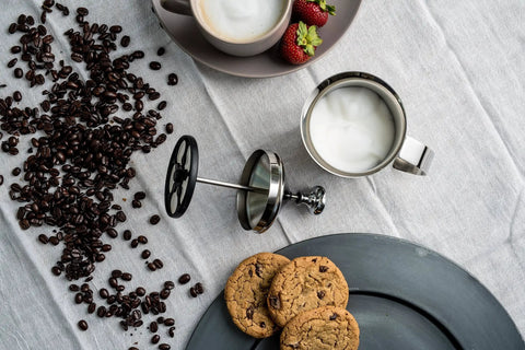 Handheld Milk Frother For Creamy Foam In Seconds, On The Go Stainless Steel Foamer beside coffee beans and cookies.