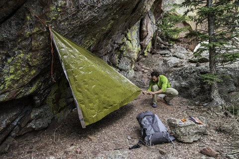 Person setting up a 90 Percent Heat Reflective Emergency Blanket Beefed Up Heavy Duty For Disaster Prep Kit as a makeshift shelter outdoors.