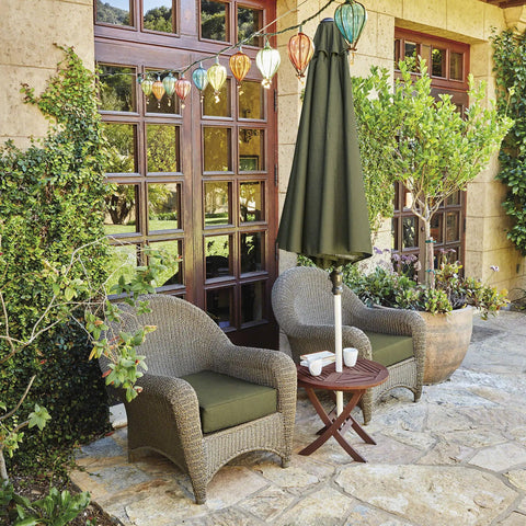 Outdoor patio seating area with two wicker chairs, green cushions, a round table, and an umbrella, surrounded by plants and lanterns.