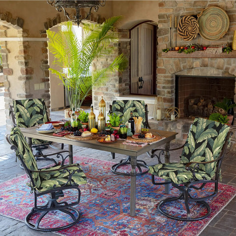Outdoor dining area with tropical print chair cushions on swivel chairs, stone fireplace, and wooden table with decorative items.