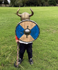 Child in Kids Viking Horned Helmet For Costume Play LARP And Halloween with shield on green grass field.