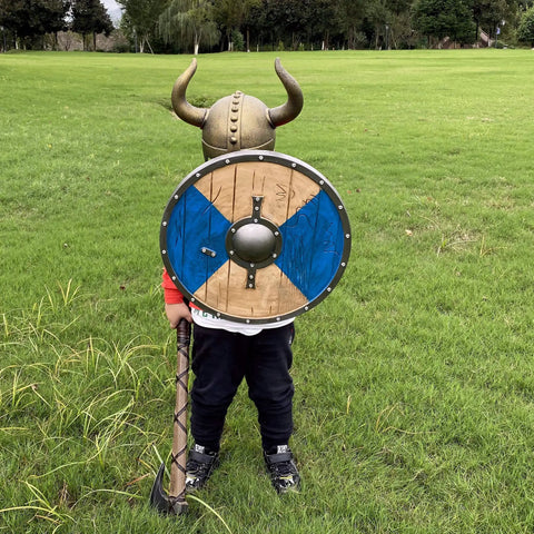 Child in Kids Viking Horned Helmet For Costume Play LARP And Halloween with shield on green grass field.