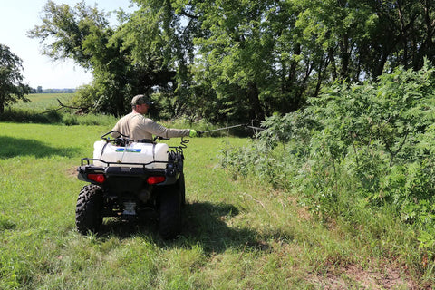Man using 15 Gallon ATV Boomless Broadcast Sprayer With 16 Foot Swath, 2.2 GPM On Demand Diaphragm Pump near trees