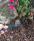 Person using Trake Garden Hand Tool Combo Trowel And Rake For Planting And Cultivating In Tight Spaces, digging near flowers.