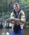 Person holding a trout near a forest stream, wearing a jacket and jeans, with fishing rod visible in the background.
