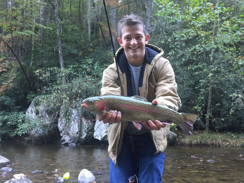Person holding a trout near a forest stream, wearing a jacket and jeans, with fishing rod visible in the background.
