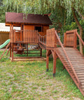 Wooden outdoor playset with slide, bridge, and small house, surrounded by grass and trees in a yard setting.
