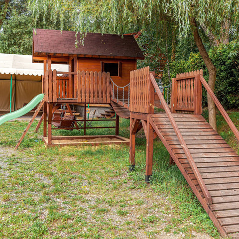 Wooden outdoor playset with slide, bridge, and small house, surrounded by grass and trees in a yard setting.