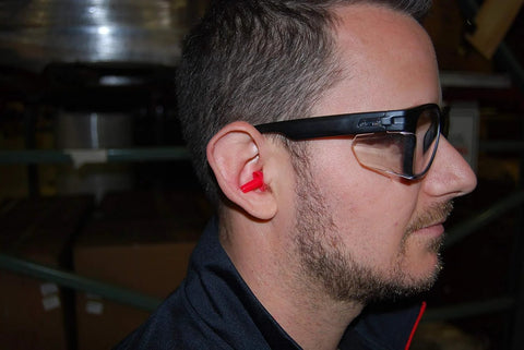 Man wearing ANSI Z87.1 Safety Glasses With Anti Fog UV Protection Clear Lens Side Shields indoors, showing side view with red earplugs.