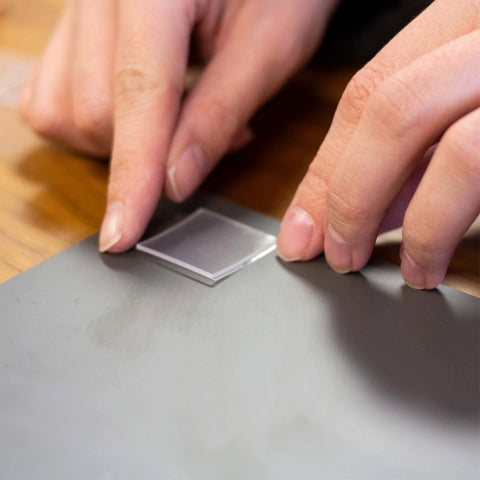 Person applying a Tough And Clear Double Sided Tape Square for Mounting, 24 Pre-Cut 1-Inch Squares on a gray surface indoors.