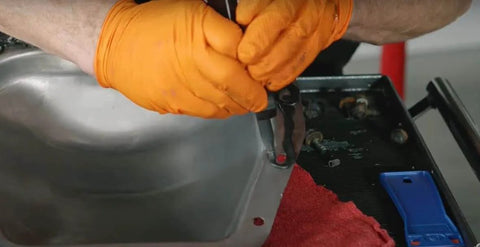 Mechanic applying Ultra Black Maximum Oil Resistant RTV Silicone Gasket Maker 9.5 Oz to metal surface with gloves.
