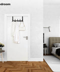 Over the Door Towel Rack with 15 Triple Hooks Black Metal in a bedroom, holding a bag and coats on a white door.