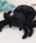 Ultra Soft Black Spider Plush Toy For Kids And Adults on beige surface, 9.8-inch cozy huggable jumping spider stuffed animal pillow.