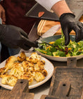 Uno chef che indossa guanti da visita in vinile nero, monouso, senza lattice e senza talco (confezione da 100), mentre prepara un'insalata e delle verdure grigliate.