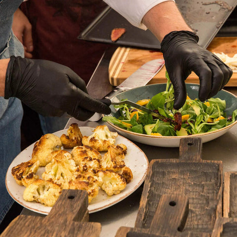 Uno chef che indossa guanti da visita in vinile nero, monouso, senza lattice e senza talco (confezione da 100), mentre prepara un'insalata e delle verdure grigliate.