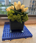 Flowers in a vase placed on a blue nestable plastic pallet. Plastic Pallets For Basement Storage And Garage Use Nestable Lightweight Waterproof Design For Basement Garage And Supermarkets Odorless Premium New PE Material 1000 Lb Heavy Duty Load Capacity Long Lasting Three Year Lifespan.