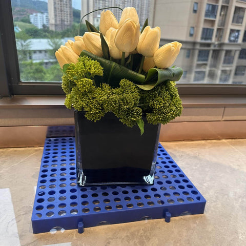 Flowers in a vase placed on a blue nestable plastic pallet. Plastic Pallets For Basement Storage And Garage Use Nestable Lightweight Waterproof Design For Basement Garage And Supermarkets Odorless Premium New PE Material 1000 Lb Heavy Duty Load Capacity Long Lasting Three Year Lifespan.