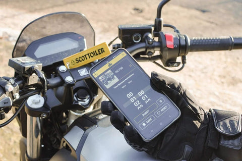 Gloved hand holding smartphone showing Micro X System 3.0 Plus Bluetooth App for motorcycle chain lubrication on bike dashboard.