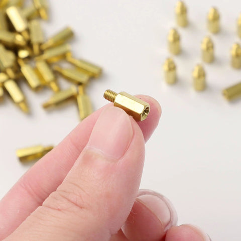 Close-up of hand holding M3 Brass Hex Standoff Spacers Set For PC Build And Motherboard 50 Pieces With Male To Female Threads 9mm And 4mm Lengths Durable PCB Hardware Kit With Nuts And Screws Uxcell showing brass material and thread size.