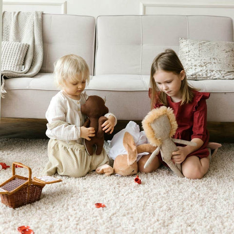 Two children playing with stuffed animals on a beige textured rug in a cozy living room setting.
