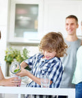 Children pouring water in a modern kitchen, with parents smiling in the background. Bright, airy home setting.