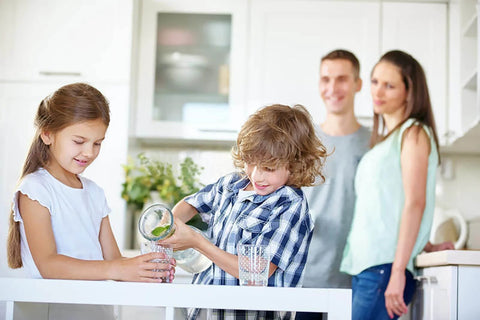 Children pouring water in a modern kitchen, with parents smiling in the background. Bright, airy home setting.