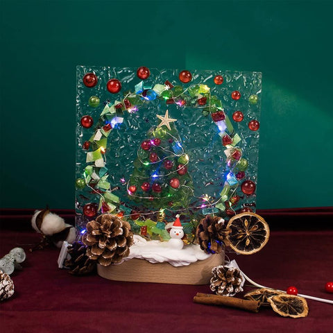 Christmas-themed decoration with pine cones, lights, and a snowman on a rustic base, set against a festive green background.