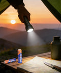 Person using flashlight powered by Combo Pack Of 24 AA And AAA Batteries 12 Each 1.5V Alkaline Heavy Duty, on a camping trip at sunset.