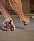 Close-up of a hand using a compact 50 ft chalk reel with blue chalk to mark a straight line on a construction site floor.