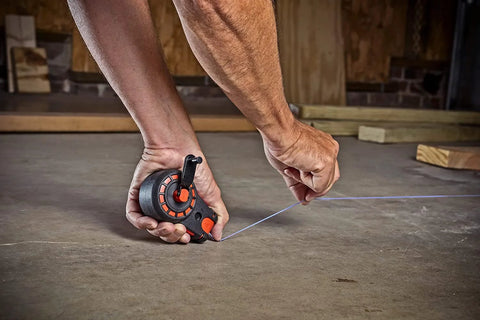 Close-up of a hand using a compact 50 ft chalk reel with blue chalk to mark a straight line on a construction site floor.