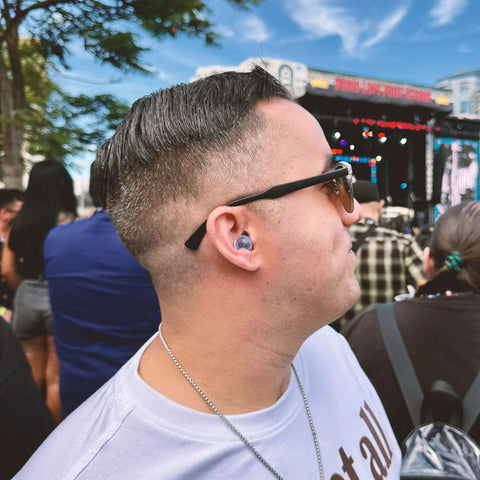Person wearing high fidelity reusable earplugs with blue filters for concerts and live music at an outdoor event.