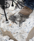 Worker using a pneumatic drill with smoothing air hammer bits set on concrete block outdoors, wearing brown work boots.