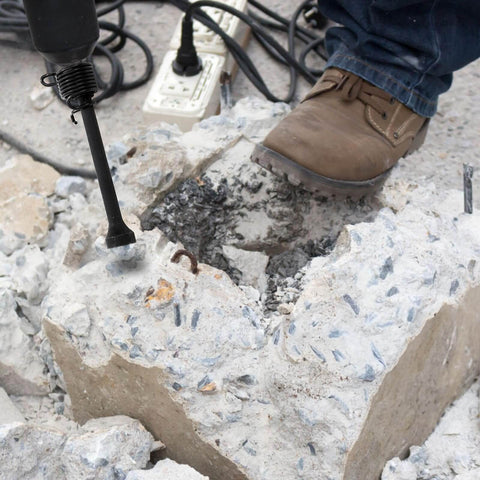 Worker using a pneumatic drill with smoothing air hammer bits set on concrete block outdoors, wearing brown work boots.