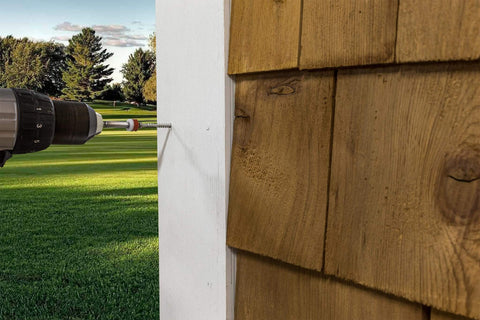 Close-up of a drill installing siding on a house exterior, with a view of a garden and the sunny outdoors in the background.