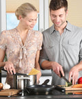 Two people cooking in a kitchen, using cutting boards and various utensils, preparing vegetables and pasta in a casual setting.