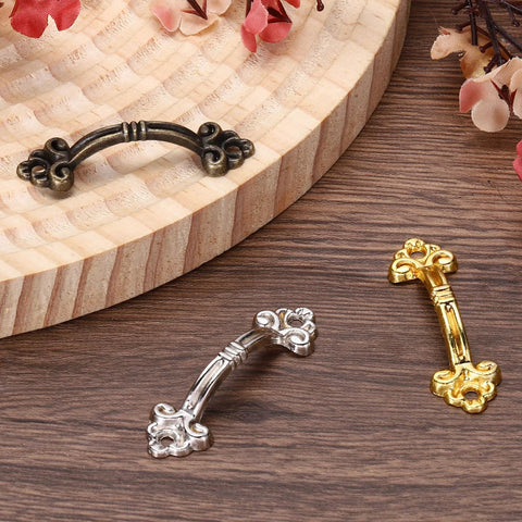 Three mini decorative drawer pulls in black, silver, and gold finishes on a wooden surface, showcasing the 20Pcs Mini Decorative Drawer Pulls Gold Finish 1.9 Inch Zinc Alloy Cabinet Handles With Screws For Furniture Dresser Jewelry Boxes And Sliding Doors Vintage Style Screw Mounted Hardware.