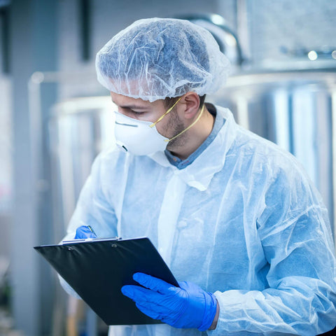 Person wearing white disposable lab coat for adults, medium size, 40 GSM polypropylene, with hair net and mask, holding a clipboard.