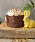 Easter bread with frosting in a brown panettone paper mold, surrounded by a yellow bunny and colorful sprinkles on a wooden board.