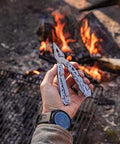 Man holding Suspension-NXT 15-In-1 EDC Multi Tool With Pocket Knife, Needle Nose Pliers And Wire Stripper by a campfire