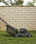 Person pushing a lawnmower on green grass in a backyard with a brick wall and trees in the background.